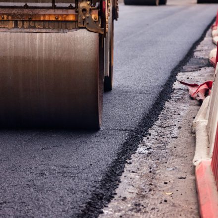 Workers are paving asphalt on a city street, with a roller compactor smoothing out the surface. Road barriers are visible, indicating an active construction zone in an urban setting.